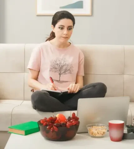 Woman taking notes in front of a laptop with a bowl of fresh fruit on the table, planning her thyroid management diet.